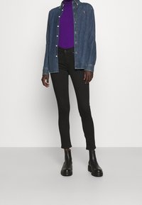 Denim shirt in dark blue with buttons, worn over a purple turtleneck. Slim-fit black pants and ankle boots complete the outfit.