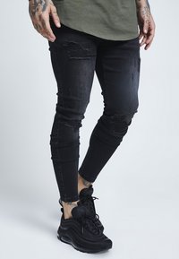 Svarta skinny jeans med slitna detaljer och fransade lappar. Bärs tillsammans med svarta sneakers, vilket visar upp en åtsittande design och mörk tvätt.