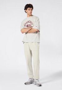 Man die met gekruiste armen staat, gekleed in een beige oversized T-shirt met de tekst "Oceancrest 1925", lichtbeige broek en grijze sneakers.