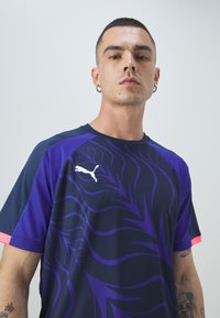 Moški s kratkimi lasmi in uhani nosi športno majico Puma v mornarsko modri in vijolični barvi z abstraktnimi vzorci ter vidnimi tetovažami na podlahti.