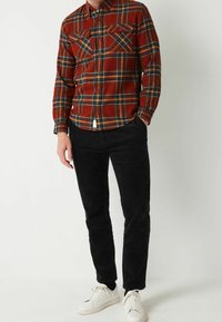 Chemise à carreaux rouge et bleue avec deux poches poitrine, associée à un pantalon en velours côtelé noir et des baskets blanches. Fond uni.