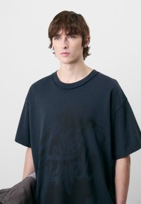 Camiseta de algodón negra de gran tamaño con un diseño gráfico texturizado. Cuenta con un cuello redondo y mangas cortas, llevada con una chaqueta gris.