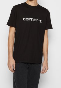 Mann in schwarzem Carhartt-T-Shirt mit weißem Logo-Text, kombiniert mit dunklen Jeans, steht vor einem schlichten hellen Hintergrund.