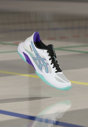 Fehér és lila sportcipő fekete fűzőkkel, egy beltéri sportpálya fölött lebeg, ahol színes vonalak láthatók.