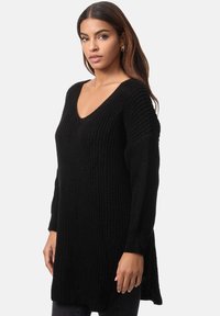Schwarzer, oversized Strickpullover mit V-Ausschnitt, langen Ärmeln, gerippter Textur, seitlichen Schlitzen und lockerer Passform, kombiniert mit dunklen Hosen.