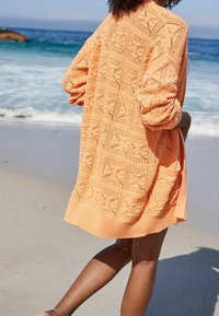 Ażurowy, brzoskwiniowy kardigan z geometrycznymi wzorami, z prążkowanymi mankietami i dołem; noszony na lekkim topie, pokazany na plaży.