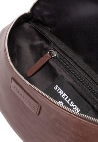 Notranjost rjave usnjene torbe z črnim podlogom, žep z zadrgo in rjavim usnjenim potegom ter etiketo z napisom "STRELLSON".