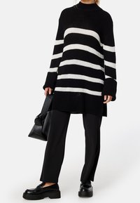 Schwarz-weiß gestreifter, übergroßer Strickpullover mit langen Ärmeln, kombiniert mit schwarzen, weiten Hosen und klobigen schwarzen Schuhen.