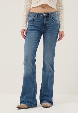 Frau trägt mittelhohe blaue Bootcut-Jeans, kombiniert mit beigen spitzen Schuhen und einem kurzen weißen Strickpullover.