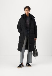 Svart pufferjacka med huva, quiltat mönster och knäppningar. Åtsidosatt med lösa denimjeans och svarta sneakers med vita ränder.