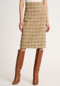 Jupe crayon en tweed beige avec un motif texturé, taille cintrée, longueur genou, associée à des bottes en cuir marron à talons hauts.