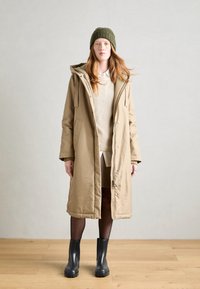 Cappotto parka beige con cappuccio, abbinato a un berretto di maglia verde con texture, indossato sopra un maglione e una gonna beige, con stivali neri.