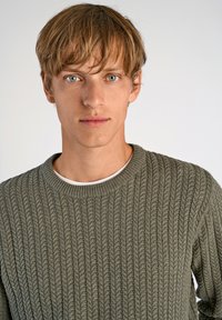 Pull en maille côtelée vert olive avec un col rond, porté sur un t-shirt blanc, présentant un motif texturé et une épaisseur moyenne.