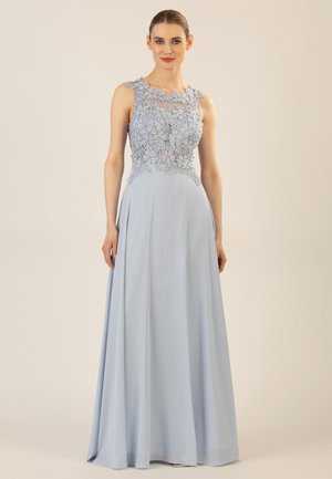 Frau steht, trägt ein ärmelloses hellblaues Kleid mit Blumen-Spitzenoberteil und fließendem Rock vor einem schlichten hellen Hintergrund.