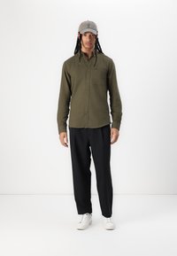 Camisa verde oliva de botones con un bolsillo en el pecho, combinada con pantalones negros y zapatillas blancas. La persona lleva una gorra gris claro.