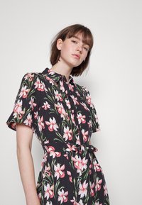 Vestido camisola floral com mangas curtas, fundo preto, padrão de flores brancas e rosas, fecho de botões e detalhe de cintura com laço.