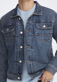 Sininen farkkutakki, jossa on klassinen muotoilu, napitettava etuosa, neljä taskua ja erottuvat ompeluyksityiskohdat. Vuorattu vaaleansinisen villapaidan yli.