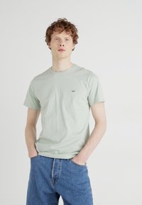 Camiseta de algodón verde claro con mangas cortas, que presenta un pequeño logotipo oscuro en azul de "Lee" en el pecho. Combinada con jeans de mezclilla azul.