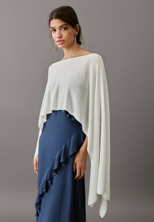Femme portant un poncho asymétrique blanc sur une robe bleu marine avec un détail de volants diagonaux, debout devant un fond gris uni.