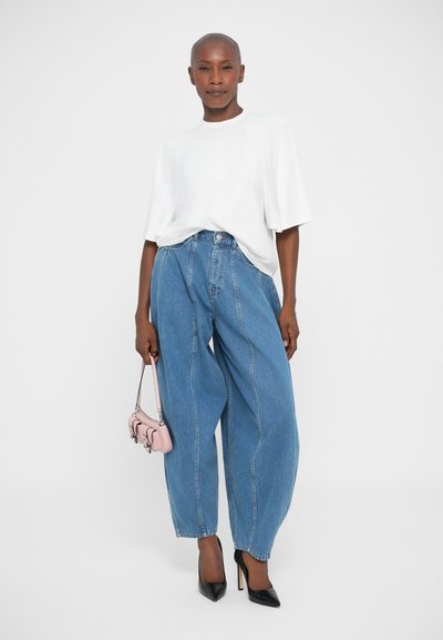 Vrouw in losse blauwe jeans, wit oversized t-shirt, zwarte hoge hakken, met een kleine roze handtas, staand tegen een effen lichte achtergrond.