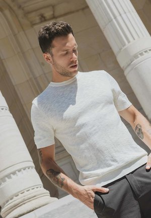 Homme aux cheveux courts et foncés avec des tatouages sur les bras, portant une chemise blanche à manches courtes et un pantalon noir, debout près de grandes colonnes de pierre.