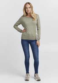 Grønn langermet genser med rund hals, kombinert med blå smale jeans og grå joggesko, med teksturert stoff og en subtil logo.