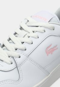 Bijeli tenisice s glatkom kožom, teksturiranim bijelim vezicama, ružičastim Lacoste logotipom i niskim siluetom.
