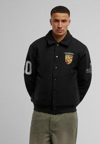 Black wool varsity jacket with a flat collar, snap buttons, and side pockets. Features a colorful emblem and "40" printed on the sleeve.