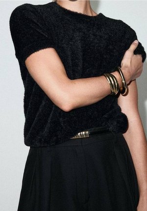 Personne portant un haut noir à manches courtes duveteux et un pantalon noir, avec des bracelets dorés au poignet gauche, bras croisés sur la poitrine.