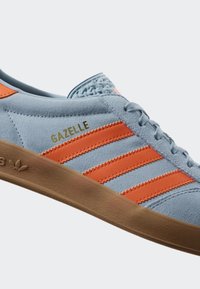 Light blue suede sneaker with orange three stripes and "Gazelle" text. Brown rubber sole, textured upper, and woven detail on the tongue.