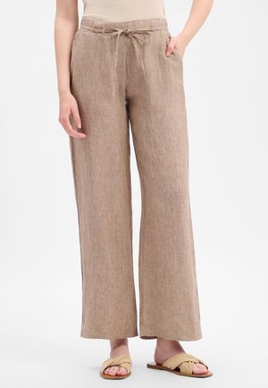 Vrouw draagt beige linnen broek met wijde pijpen en een trekkoord in de taille, en bruine instapsandalen, staand tegen een witte achtergrond.