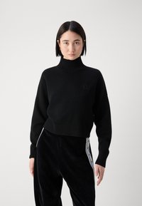 Zwarte gebreide trui met een hoge col en geribde mouwen, met een klein logo op de borst. Gecombineerd met zwarte fluwelen broek met een zijstreep.
