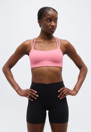 Atletische vrouw met gevlochten haar, gekleed in een roze sportbeha en zwarte shorts met hoge taille, staand met handen in de zij en zijwaarts kijkend.