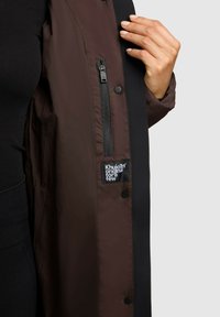 Giacca antivento marrone e nera con tasca con cerniera e etichetta in tessuto. Texture liscia con bottoni automatici lungo l'apertura frontale.