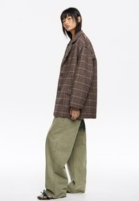 Blazer de cuadro marrón de gran tamaño con botones de doble botonadura, combinado con pantalones anchos de color verde oliva y sandalias multicolor.