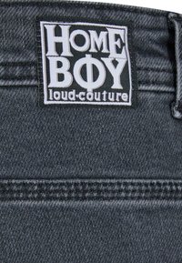 Tissu en denim noir avec une surface texturée arborant une étiquette blanche proéminente indiquant "HOME BOY loud-couture." Coutures propres et cousues.
