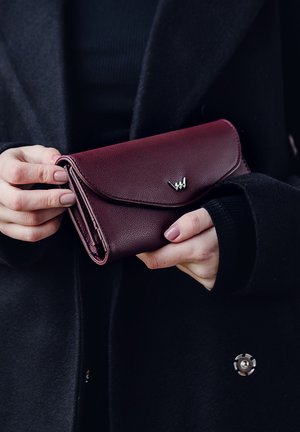Burgundy leather wallet with a textured finish, curved flap closure, and a silver logo accent, held in hands against a black coat.