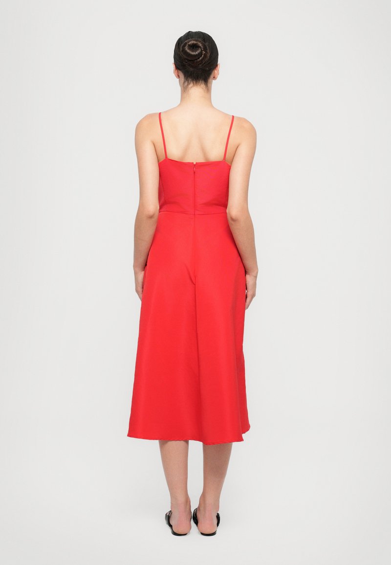 Une femme avec un chignon porte une robe rouge sans manches, vue de dos, sur un fond neutre.