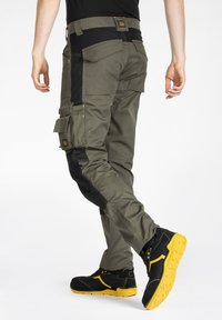 Pantalons cargo en vert olive avec des accents noirs, dotés de plusieurs poches, d'une coupe ajustée, portés avec des chaussures noires à semelles jaunes.