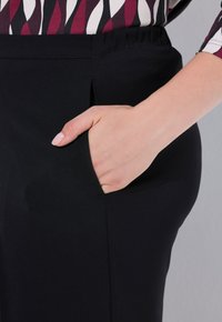 Zwarte broek met een zijzak, met een gladde textuur en een ontspannen tailleband. Het ontwerp bevat een subtiele plooi-detail.