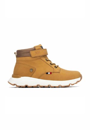 Zapatilla de caña alta color beige con suela blanca, cuello acolchado marrón, cierre de velcro, cordones y pequeño detalle de bandera roja, blanca y azul en el lateral.