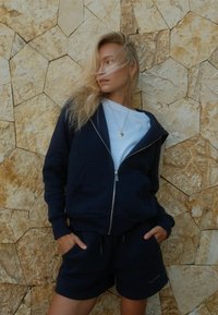 Marineblå hettegenser med glidelås og lommer, kombinert med matchende shorts. Glatt stoff med løs passform og liten logo på shortsen.