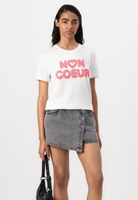 Balta medvilninė marškinėliai su rožiniu tekstu "MON ❤️ COEUR" ir pilki džinsiniai sijonai su įstrižine iškirpte ir priekiniais mygtukais. Modelis laiko juodą rankinę.