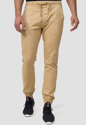 Beige joggerbukser med elastisk talje og manchetter, der har sidelommer og en blød, struktureret stof. Parret med sorte sneakers.