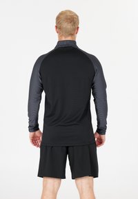 Man med kort blont hår iförd en långärmad svart och mörkgrå sporttröja och svarta shorts, vänd bort från kameran mot en vit bakgrund.