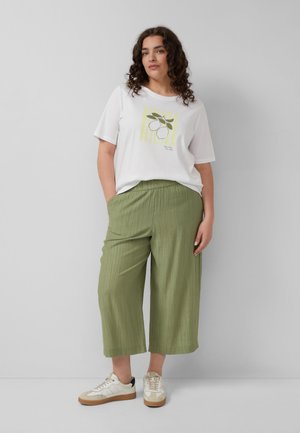 Kobieta o kręconych ciemnych włosach, ubrana w białą koszulkę z grafiką cytryny, jasnooliwkowe szerokie krótkie spodnie i białe sportowe buty.