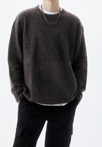 Maglione fuzzy marrone con superficie a maglia testurizzata, vestibilità rilassata, spalle basse e scollatura tonda, indossato sopra una camicia bianca.