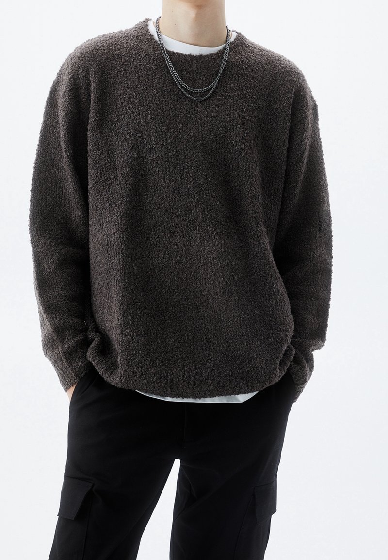 Maglione fuzzy marrone con superficie a maglia testurizzata, vestibilità rilassata, spalle basse e scollatura tonda, indossato sopra una camicia bianca.