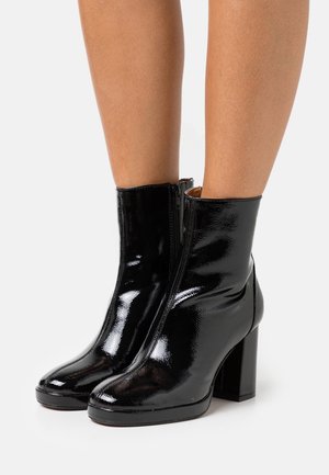Lesklé černé kotníkové boty z lakované kůže s širokým podpatkem, nošené na holých nohou na bílém pozadí.