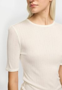 Kermakuvioinen lyhythihainen toppi, jossa on hienovarainen pistekuvio ja hapsureunus. Pehmeä tekstuuri, vartalonmyötäinen malli. Läheltä otettu kuva kauluksesta ja olkapäästä.